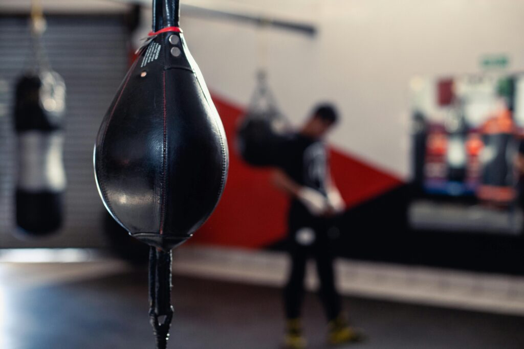 Focused view of a punching bag in a gym setting with an out-of-focus boxer in the background.